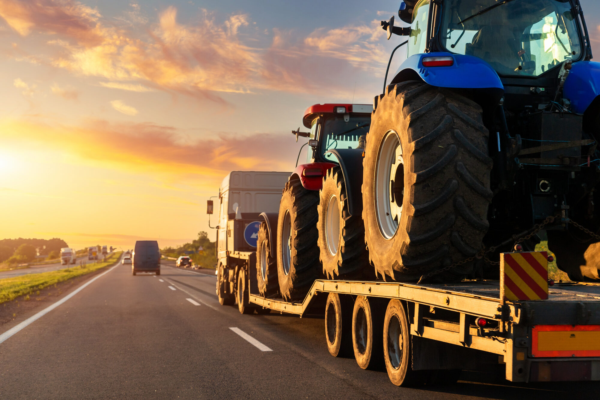 POV heavy industrial truck semi trailer flatbed platform transport two big modern farming tractor machine on common highway road at sunset sunrise sky. Agricultural equipment transportation service